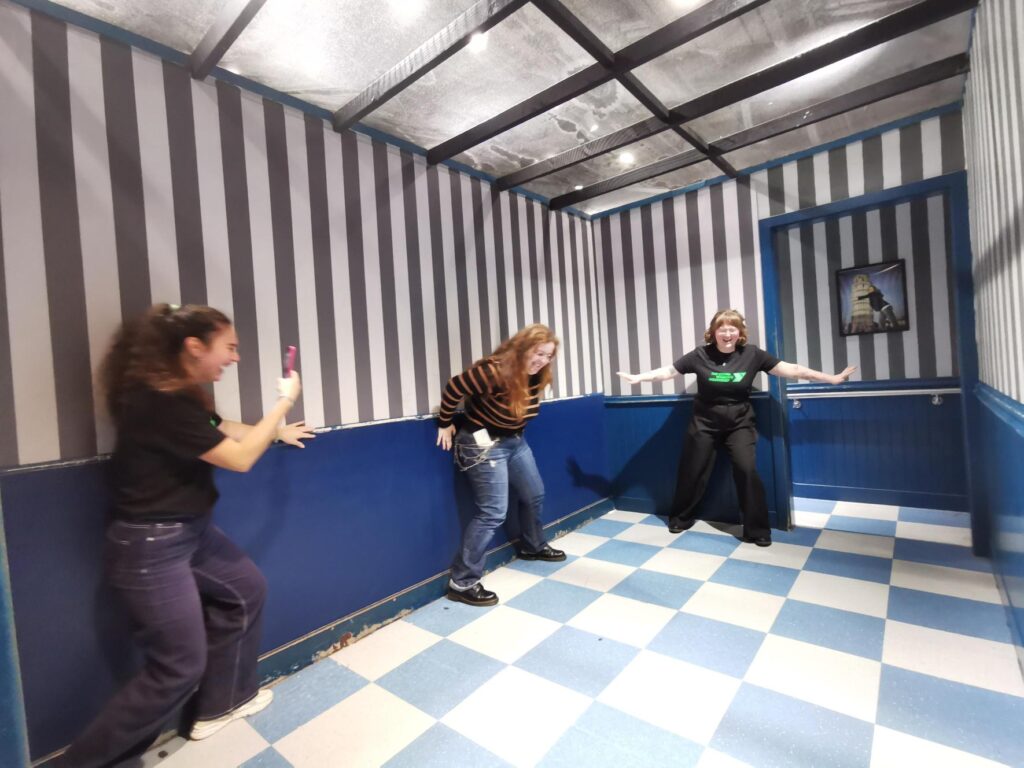 Three young women are in an optical illusion which makes some of them look smaller. They're all laughing.