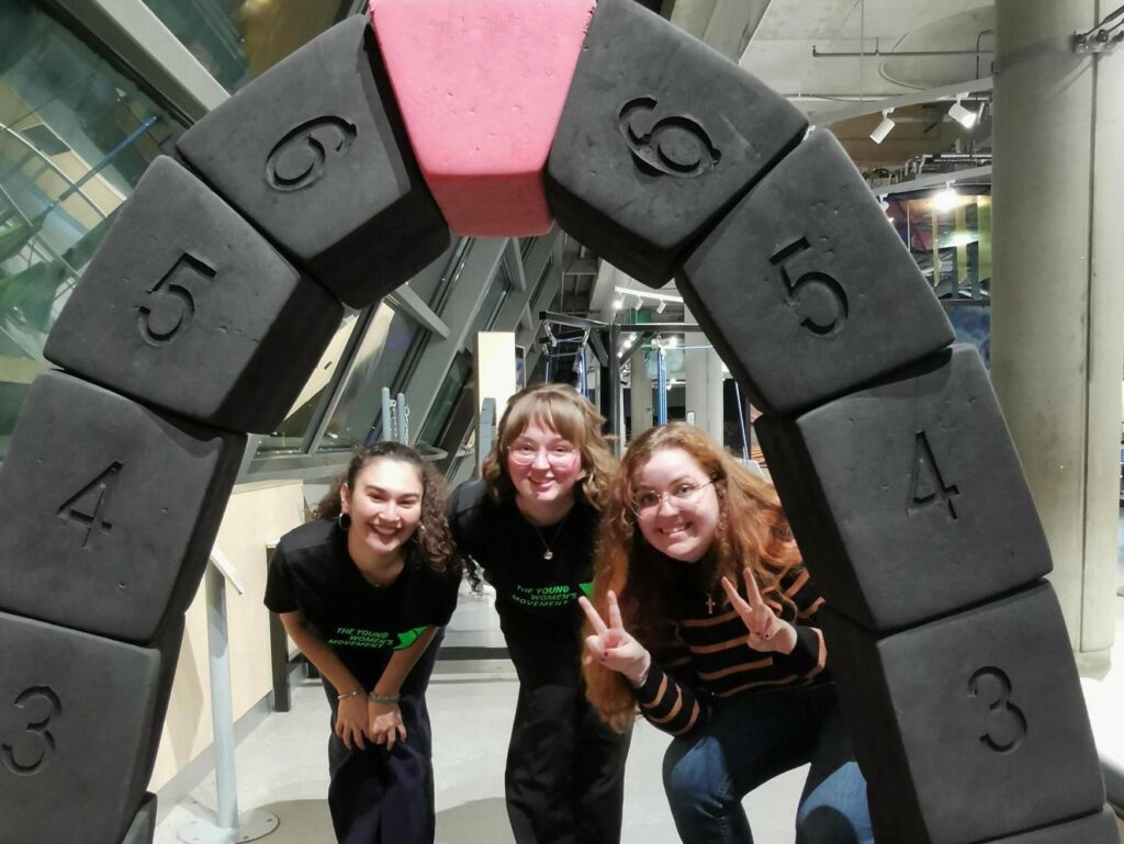 Three young women are smiling underneath an archway made of black numbered blocks. They have pale skin, and two of them are wearing The Young Women's Movement t-shirts.