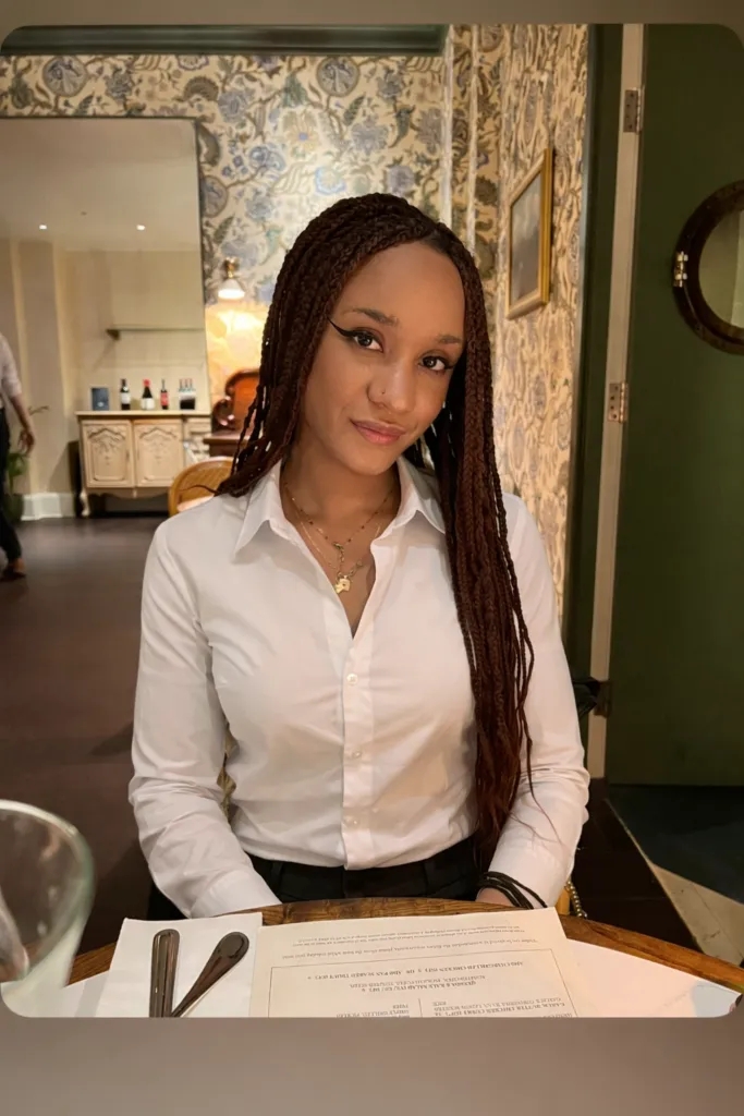 Stella has dark skin and long hair in braids. She is wearing a white shirt and sitting in a restaurant.