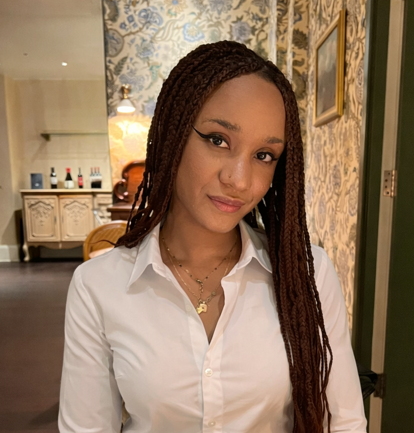 Stella has dark skin and long hair in braids. She is wearing a white shirt and sitting in a restaurant.