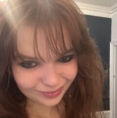 Emma has pale skin and red hair with a fringe. She's smiling at the camera.