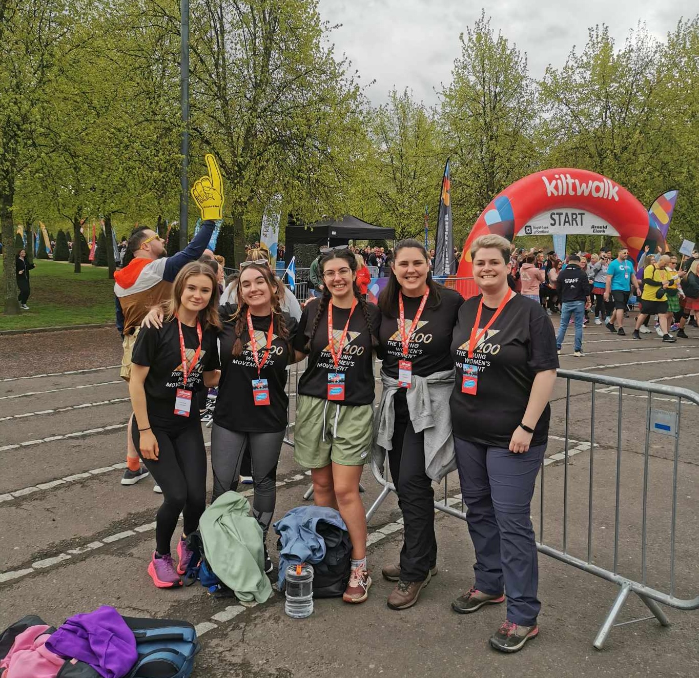 A group of five women stand in front of a kiltwalk banner. They are all wearing young women's movement t-shirts.