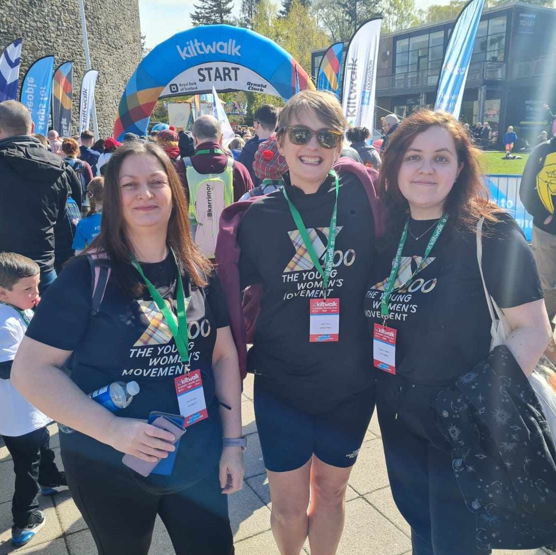 Three women smile at the camera. Behind them is the kiltwalk finish line.
