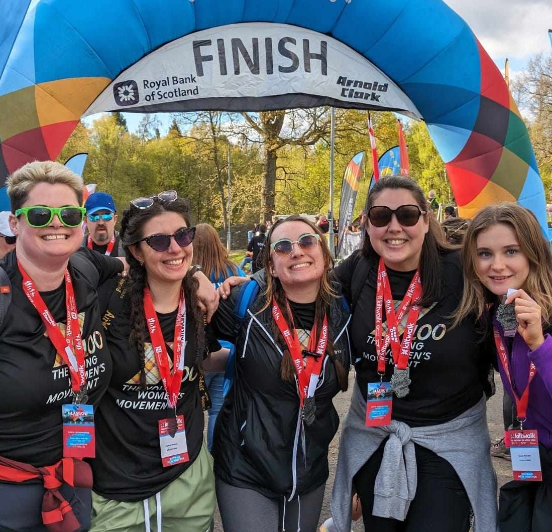 Five women smile at the camera. behind them is the kiltwalk finish line.