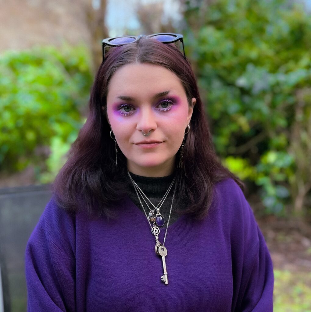 Alba has pale skin and dark hair. She is wearing a purple jumper and purple eyshadow
