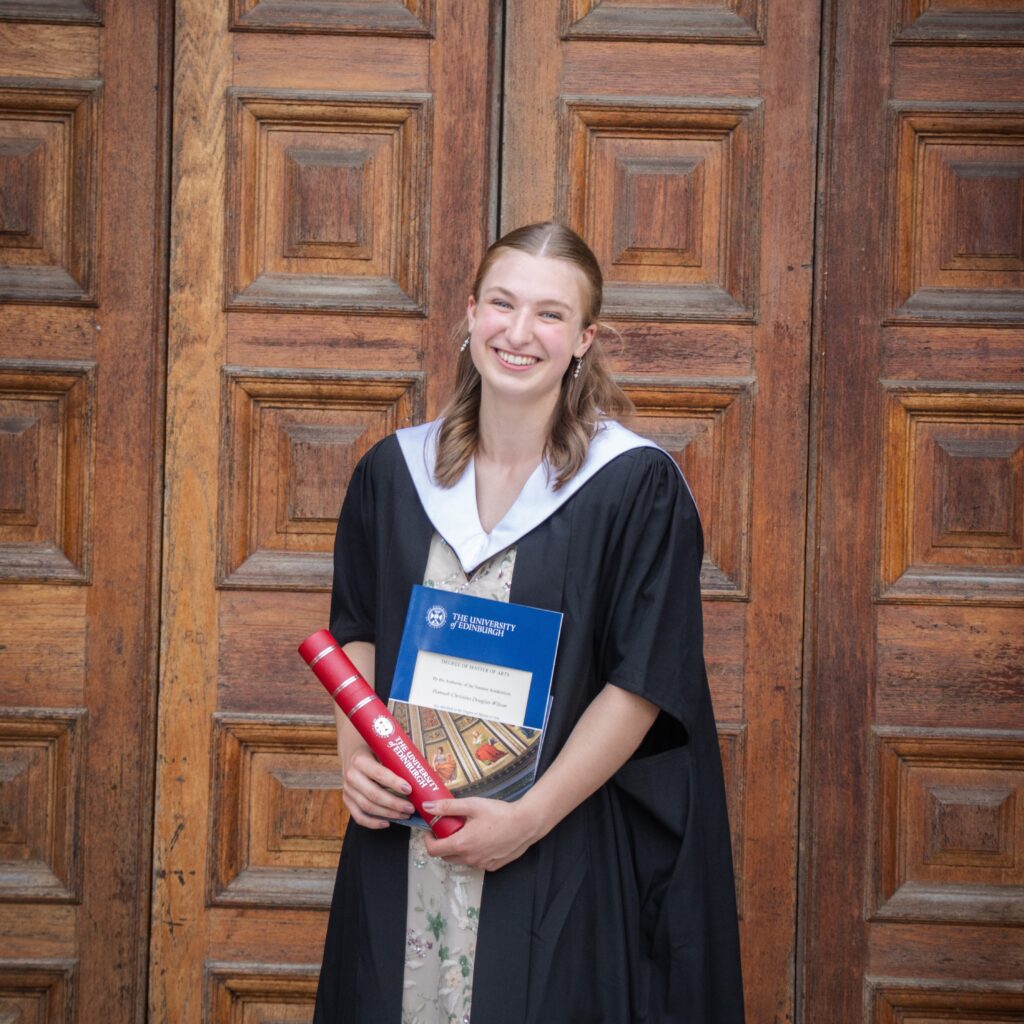 Hannah has pale skin and light hair. She is wearing a graduation gown and standing in front of McEwan Hall in Edinburgh