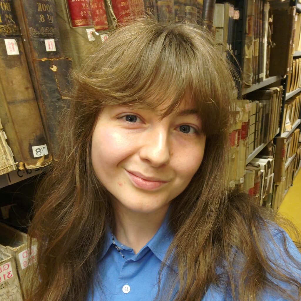 Iona has pale skin and brown hair with a fringe. She is in an archive and wearing a blue shirt.