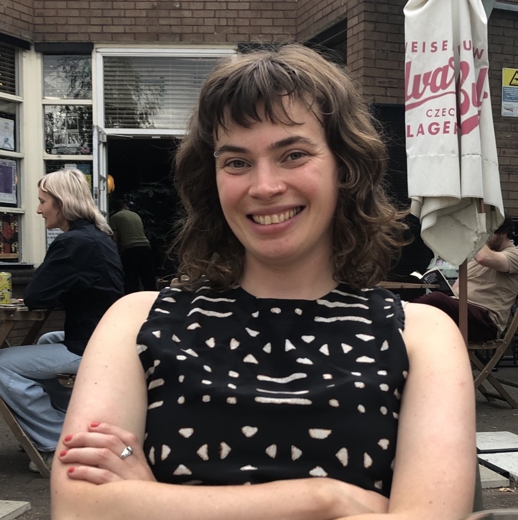 Jennie has pale skin and short dark hair with a fringe. She is smiling at the camera with her arms crossed.