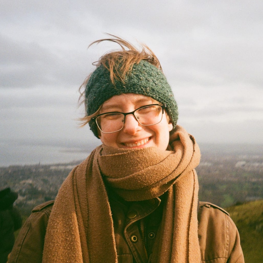 Julia has pale skin and light hair that is hidden by a knitted headband. She is wearing a scarf and glasses and is on top of a hill.