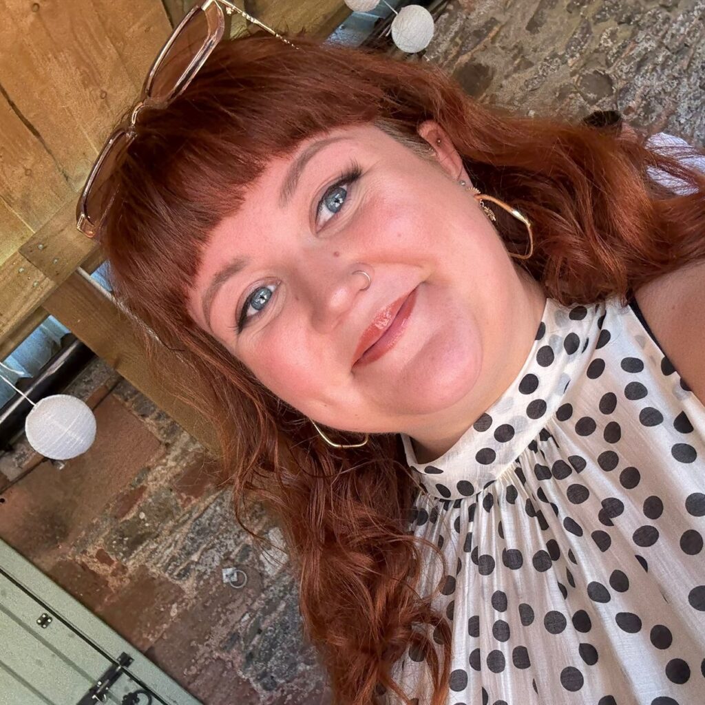 Kelly has pale skin and long red hair with a fringe. She is smiling at the camera and wearing sunglasses on her head.
