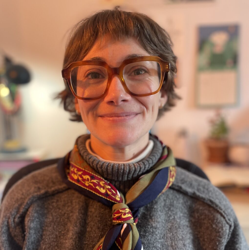 Laura has pale skin and short dark hair. She is wearing large orange glasses and smiling at the camera.