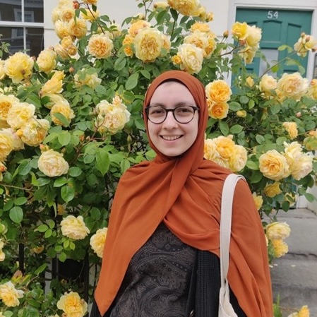 Sofia has pale skin and is wearing an orange hijab and glasses. She is smiling and standing in front of a yellow rose bush