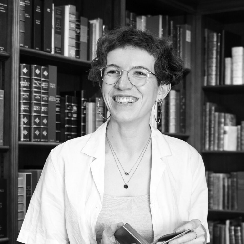 Black and white image of Suzanna, who has pale skin and short dark hair. Suzanna is standing in a library holding a book and smiling.