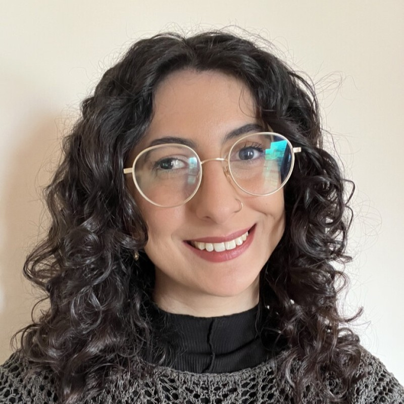 Samar has dark curly hair and glasses. She is smiling at the camera.